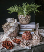 Bogata kompozycja dekoracyjna w stylu boho. Na wzorzystym materiale ze złotym nadrukiem stoją dwa ceramiczne świeczniki-karczochy – jeden brązowy, drugi beżowy z nadpaloną świecą. Przed nimi leżą trzy ceramiczne kwiaty w podobnej kolorystyce. W tle widać stos książek (jedna z tytułem "BOHEMIAN STYLE"), na którym stoi beżowa osłonka z motywem liści, zawierająca zieloną paproć. Obok znajduje się ceramiczny wazon przewiązany dekoracyjną wstążką.