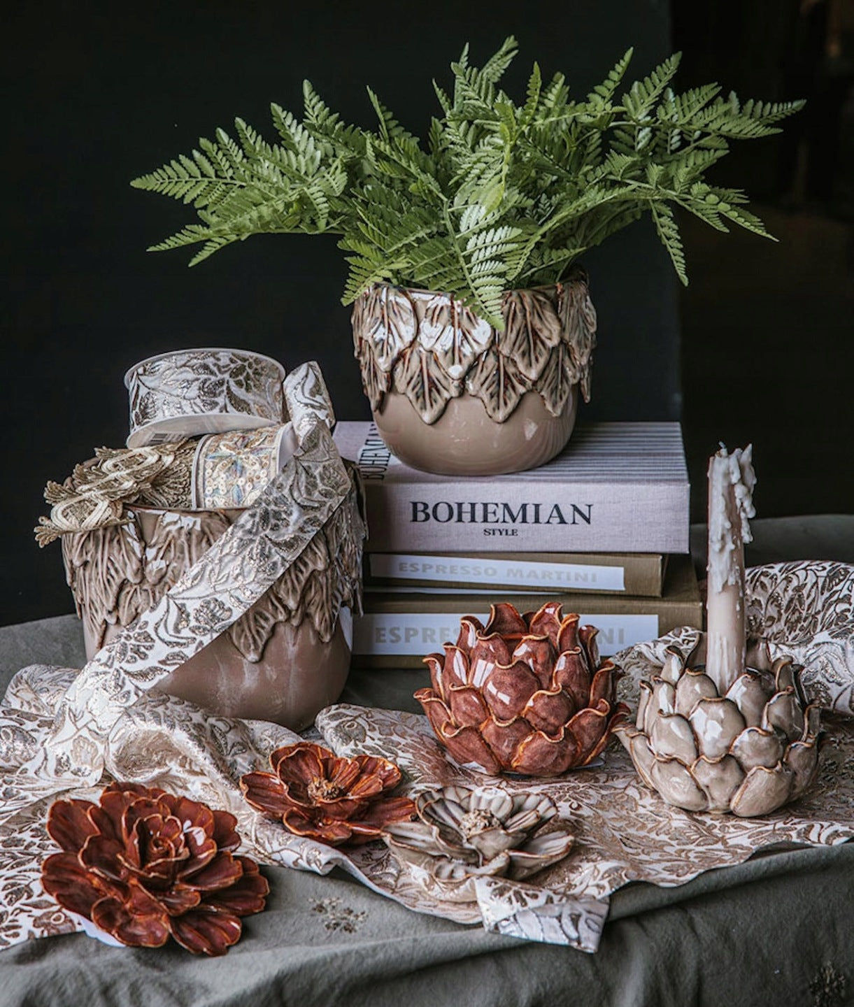Bogata kompozycja dekoracyjna w stylu boho. Na wzorzystym materiale ze złotym nadrukiem stoją dwa ceramiczne świeczniki-karczochy – jeden brązowy, drugi beżowy z nadpaloną świecą. Przed nimi leżą trzy ceramiczne kwiaty w podobnej kolorystyce. W tle widać stos książek (jedna z tytułem "BOHEMIAN STYLE"), na którym stoi beżowa osłonka z motywem liści, zawierająca zieloną paproć. Obok znajduje się ceramiczny wazon przewiązany dekoracyjną wstążką.