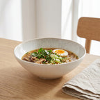 Minimalistyczna, nowoczesna kompozycja kulinarna inspirowana kuchnią azjatycką. W jasnej, głębokiej misce z efektem szkliwa reaktywnego zaserwowano aromatyczny ramen z makaronem, grzybami, świeżą kolendrą, szczypiorkiem i połówką ugotowanego jajka. Proste, drewniane tło, naturalna, lniana serwetka oraz krzesło w stylu skandynawskim idealnie podkreślają bezpretensjonalny charakter naczynia