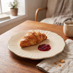 Przytulna, poranna aranżacja. Na drewnianym stole leży beżowy, ceramiczny talerz o falistym brzegu, na którym podano chrupiącego rogalika (croissant) z porcją czerwonej konfitury. Talerz spoczywa częściowo na jasnej, lnianej serwetce. W rozmytym tle widać parującą kawę w kubku, drewniany fotel oraz światło wpadające przez okno.