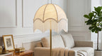 Włączona lampa podłogowa z białym, plisowanym abażurem ozdobionym frędzlami, zamocowana na prostej, mosiężnej podstawie. Lampa stoi w eleganckim, jasnym salonie tuż obok beżowej sofy. Po lewej stronie znajduje się drewniany stolik ze złotą ramką i szkatułką, a w tle po prawej rośnie wysoka roślina doniczkowa (figowiec).