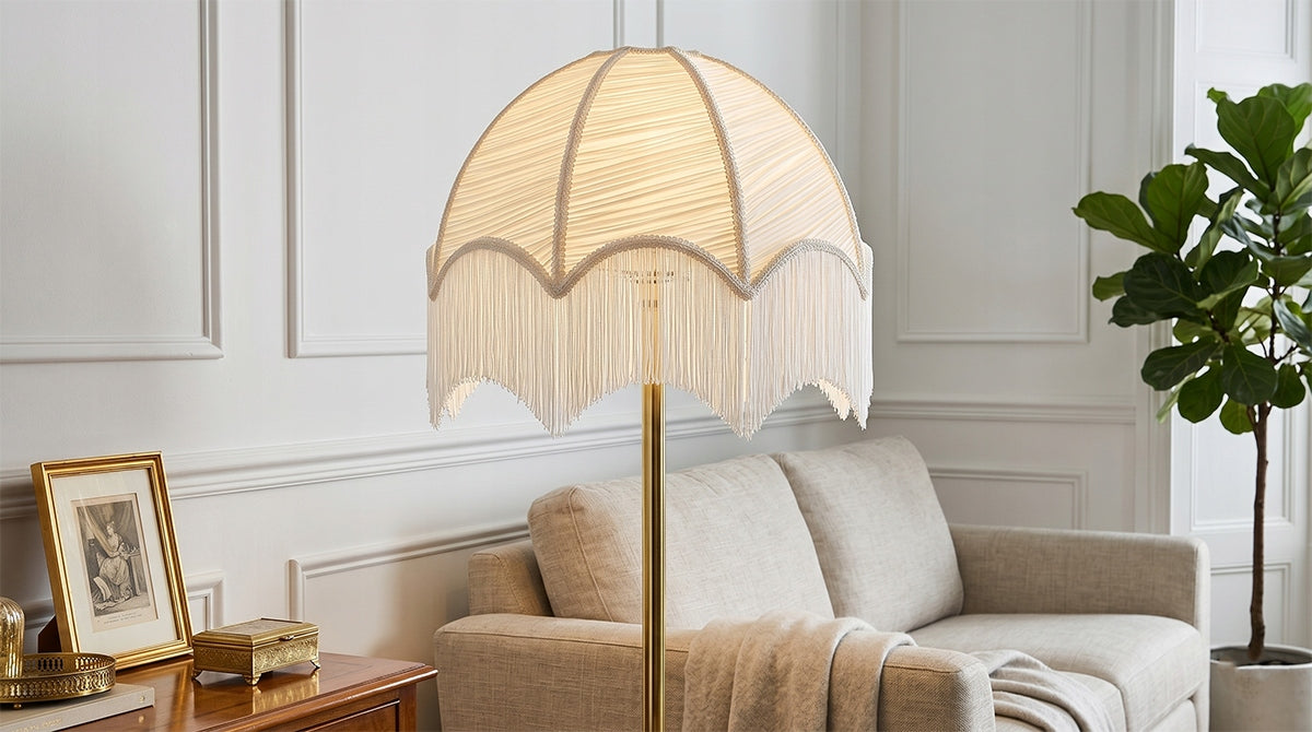 Włączona lampa podłogowa z białym, plisowanym abażurem ozdobionym frędzlami, zamocowana na prostej, mosiężnej podstawie. Lampa stoi w eleganckim, jasnym salonie tuż obok beżowej sofy. Po lewej stronie znajduje się drewniany stolik ze złotą ramką i szkatułką, a w tle po prawej rośnie wysoka roślina doniczkowa (figowiec).