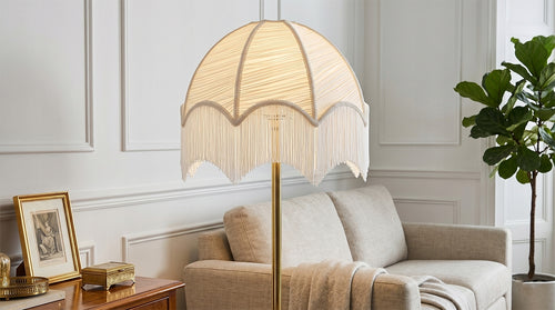 Włączona lampa podłogowa z białym, plisowanym abażurem ozdobionym frędzlami, zamocowana na prostej, mosiężnej podstawie. Lampa stoi w eleganckim, jasnym salonie tuż obok beżowej sofy. Po lewej stronie znajduje się drewniany stolik ze złotą ramką i szkatułką, a w tle po prawej rośnie wysoka roślina doniczkowa (figowiec).