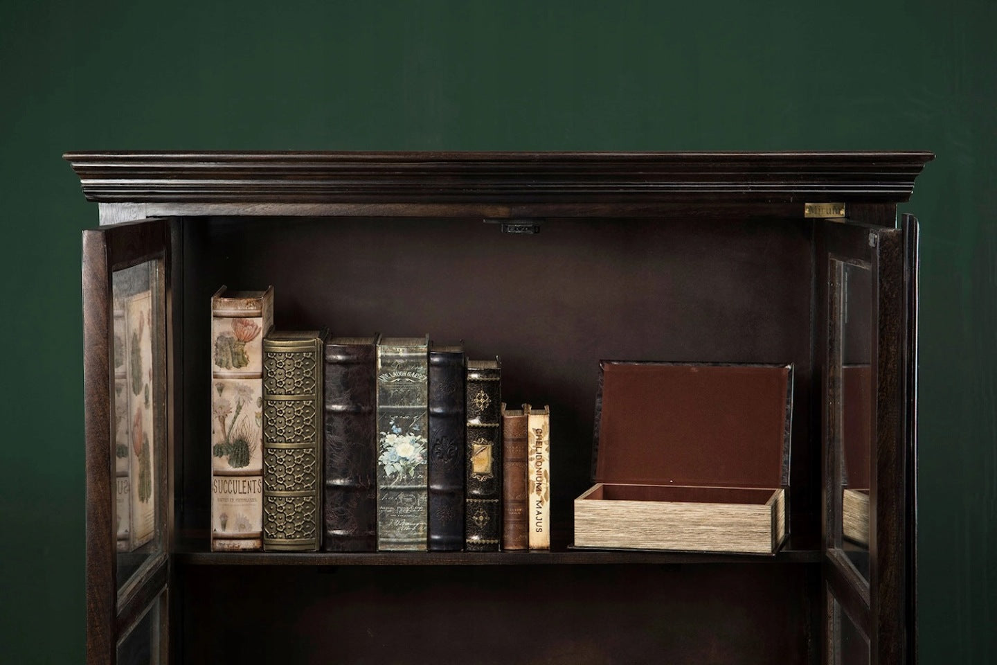 Fragment ciemnodrewnianej, otwartej biblioteczki lub witryny na tle ciemnozielonej ściany. Na półce stoi rządek dekoracyjnych pudełek imitujących stare książki o różnych okładkach (m.in. motywy roślinne, tłoczenia, imitacja skóry). Po prawej stronie leży otwarte, prostokątne pudełko obite od wewnątrz brązowym materiałem, przypominające puste opakowanie po jednej z tych "książek".