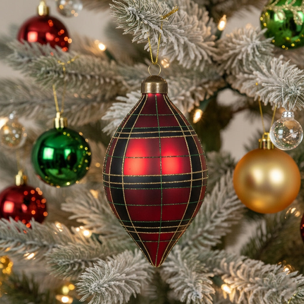 Zbliżenie na podłużną bombkę w kształcie łezki z eleganckim motywem czerwono-czarnej kraty tartan. Ozdoba wisi w centralnym punkcie oszronionej choinki. W tle widoczne są inne tradycyjne, okrągłe bombki w kolorze czerwonym, zielonym i złotym oraz delikatne, ciepłe światełka choinkowe.