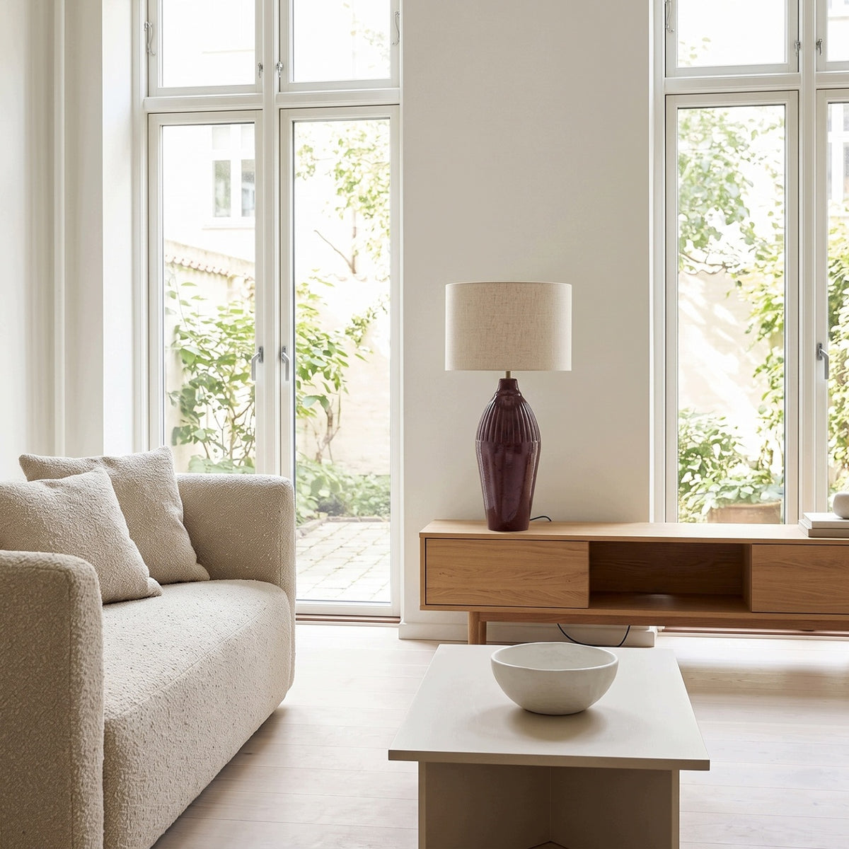 Przestronny, jasny salon urządzony w minimalistycznym, skandynawskim stylu. Bordowa lampa ceramiczna z jasnym abażurem stanowi wyrazisty akcent kolorystyczny, stojąc na podłużnej, dębowej szafce RTV. Wnętrze oświetlają wysokie, białe okna z widokiem na zielony dziedziniec. Na pierwszym planie widoczny jest nowoczesny stolik kawowy z misą oraz fragment jasnej kanapy obitej strukturalną tkaniną typu bouclé.