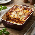 Świeżo upieczona, gorąca lasagne z widocznym, ciągnącym się, roztopionym serem na wierzchu i grubą warstwą sosu bolońskiego. Zaserwowana bezpośrednio w eleganckim, bordowym naczyniu ceramicznym (brytfannie). Naczynie stoi na drewnianym stole, a kompozycję dopełniają świeże liście bazylii, lniana serwetka oraz sztućce (nóż i widelec).