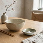 Spokojna, minimalistyczna kompozycja w stylu Japandi na jasnym, drewnianym stole. Mała, zielonkawa miseczka ceramiczna o nieregularnym kształcie, wypełniona gruboziarnistą solą morską, stoi obok większej, beżowej misy. Aranżację uzupełniają drewniane pałeczki, lniana serwetka oraz surowy wazonik z kwitnącą gałązką wiśni