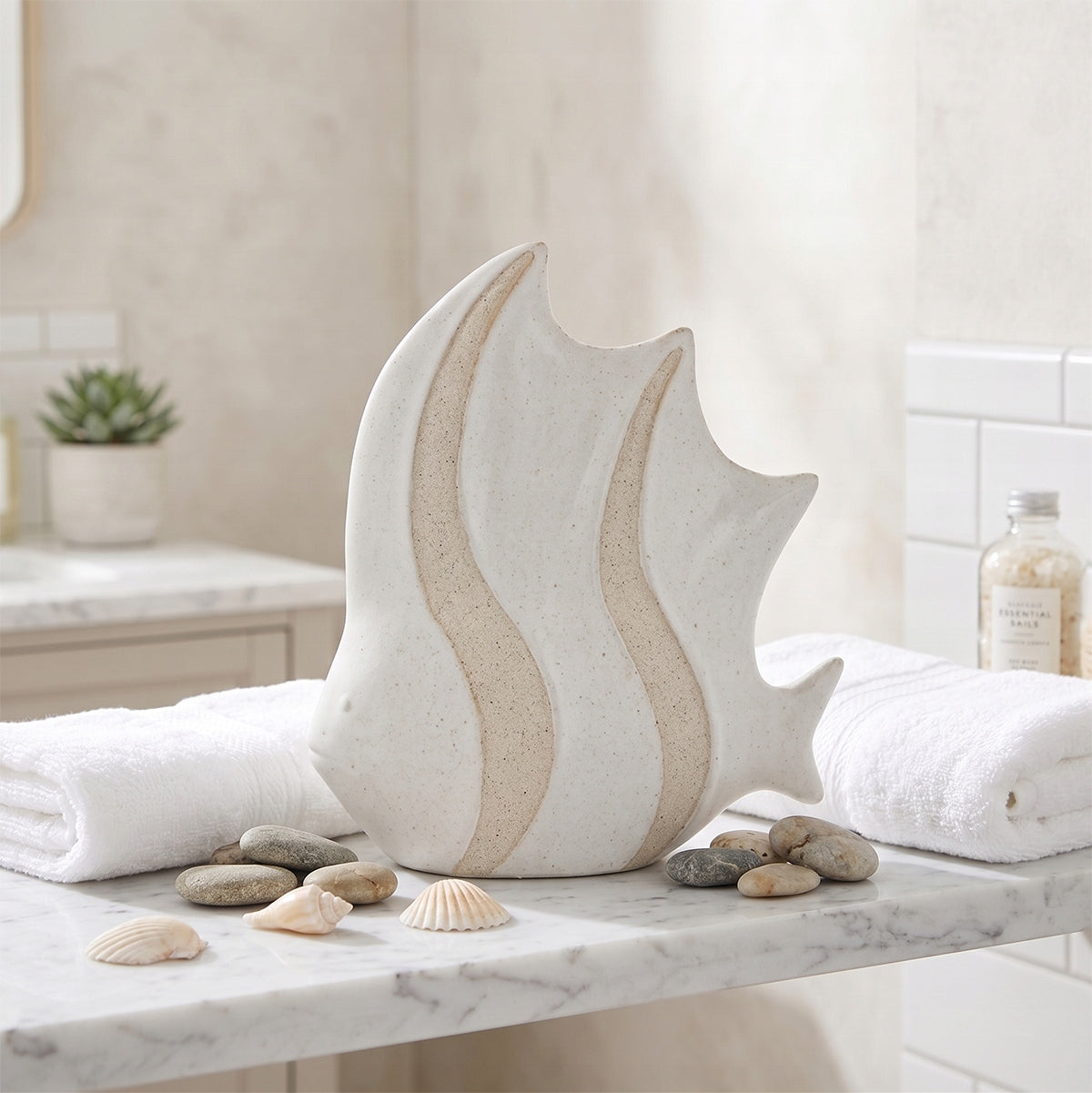 Biała, ceramiczna figurka ryby o płaskim kształcie i oryginalnej fakturze, łączącej gładkie i piaskowe pasy. Ozdoba stoi na marmurowym blacie łazienkowym, w otoczeniu naturalnych kamieni (otoczaków) i małych muszelek. Obok leży czysty, zrolowany, biały ręcznik. Wnętrze jest jasne i nawiązuje do relaksującego klimatu SPA.