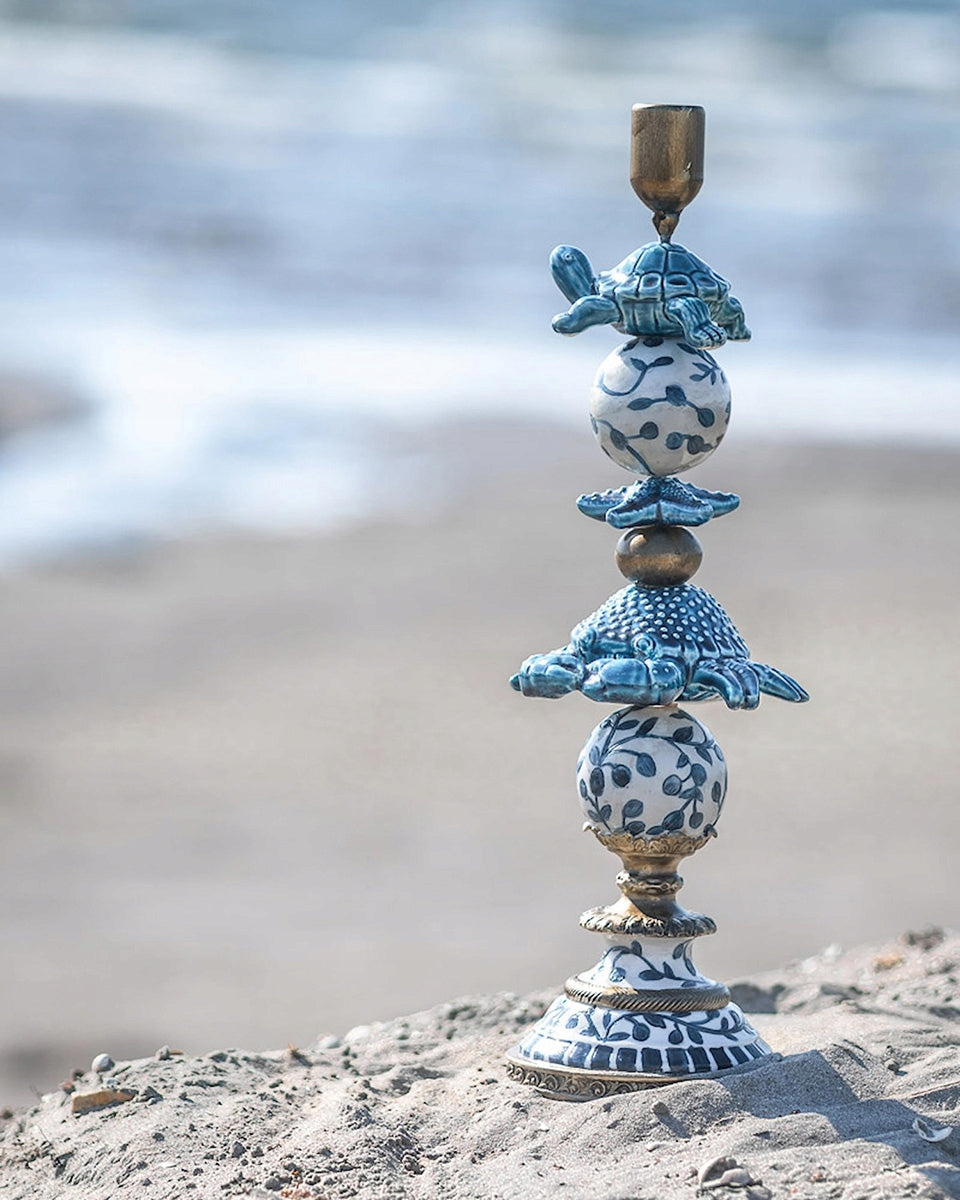 Dekoracyjny, piętrowy świecznik z morskimi motywami (krab, żółw, rozgwiazda) wkopany delikatnie w piasek na plaży. Pionowa, ceramiczno-mosiężna kompozycja idealnie wpisuje się w nadmorski krajobraz. W miękko rozmytym tle widać fale spokojnego oceanu i jasne światło słoneczne.
