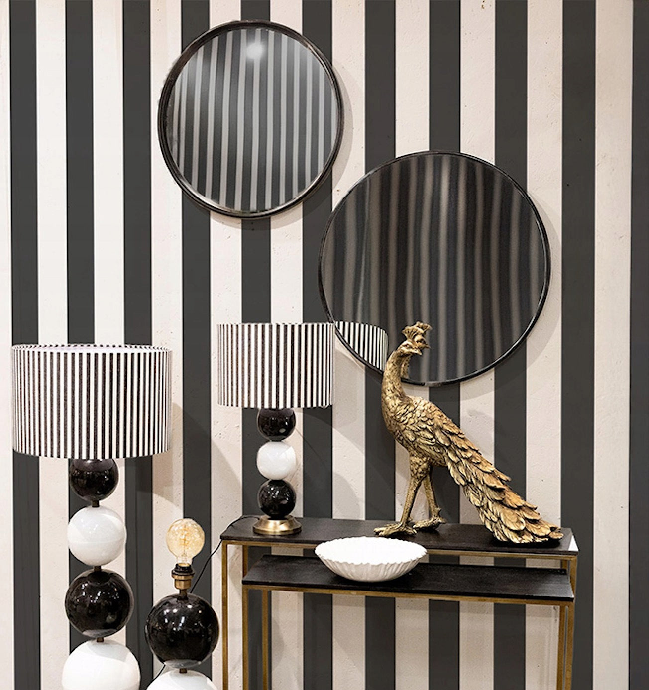 Odważna aranżacja wnętrza w stylu eklektycznym lub modern glamour. Ścianę pokrywa tapeta w mocno kontrastujące, szerokie czarno-białe pionowe pasy. Na jej tle zawieszono asymetrycznie dwa okrągłe lustra w czarnych ramach. Poniżej stoi metalowa konsola, a na niej dekoracyjna, złota figurka pawia, biała patera oraz wysokie lampy stołowe zbudowane z czarno-białych kul, zwieńczone abażurami w paski.