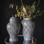 Elegancka kompozycja na czarnym tle. Na okrągłym stoliku z lustrzanym blatem stoją dwa pasujące do siebie ceramiczne naczynia: zamykana waza (ginger jar) z ciemną pokrywką oraz wysoki wazon. Oba przedmioty posiadają białe tło z gęstym, czarnym wzorem układającym się w kształt liści lub łusek. W wazonie umieszczono kompozycję z zielonego eukaliptusa oraz suszonych, ozdobnych traw, które kontrastują z mrocznym tłem. Naczynia pięknie odbijają się w lustrze blatu.