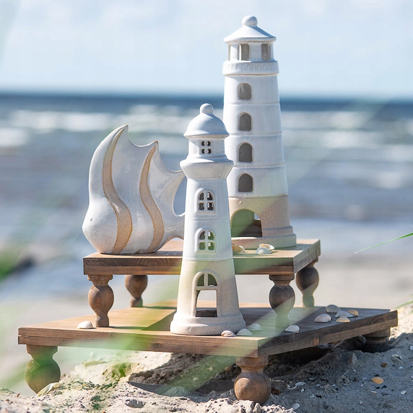Kompozycja ceramicznych ozdób w nadmorskim stylu, ustawionych na dwóch małych, drewnianych stołeczkach ułożonych kaskadowo bezpośrednio na piasku. W zestawie znajduje się figurka ryby w białym kolorze z piaskowymi pasami oraz dwie urocze latarnie morskie w podobnej, jasnej kolorystyce. W tle widać rozmyte fale morza