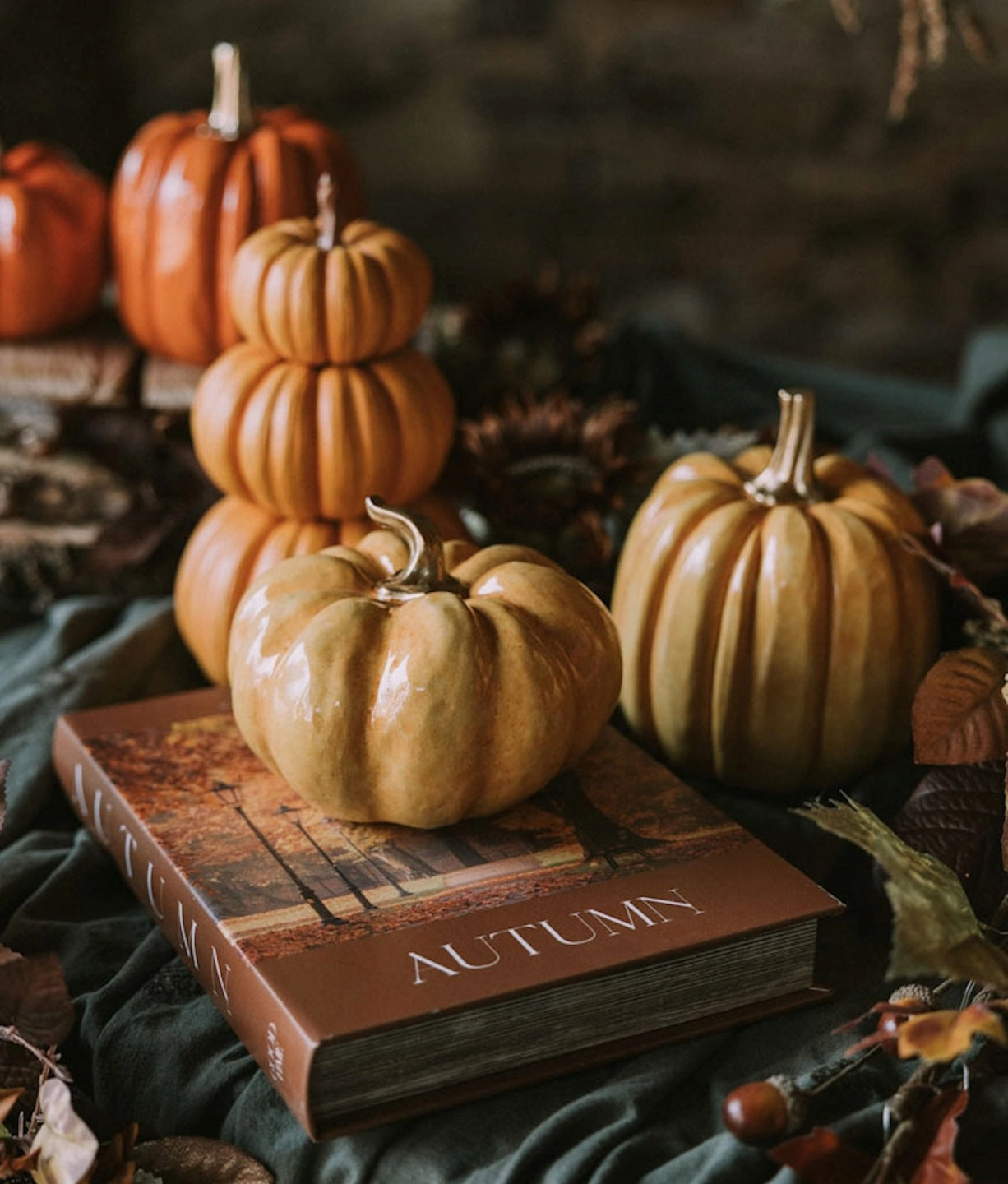 Jesienna kompozycja w ciemnych, nastrojowych barwach. Na stole przykrytym pogniecionym, ciemnozielonym materiałem leży atrapa książki z napisem "AUTUMN" i zdjęciem jesiennego parku. Na książce oraz wokół niej ułożone są ozdobne, ceramiczne dynie w różnych rozmiarach, o musztardowym i pomarańczowym wykończeniu z błyszczącym szkliwem. W tle widoczne są suszone jesienne liście i żołędzie.