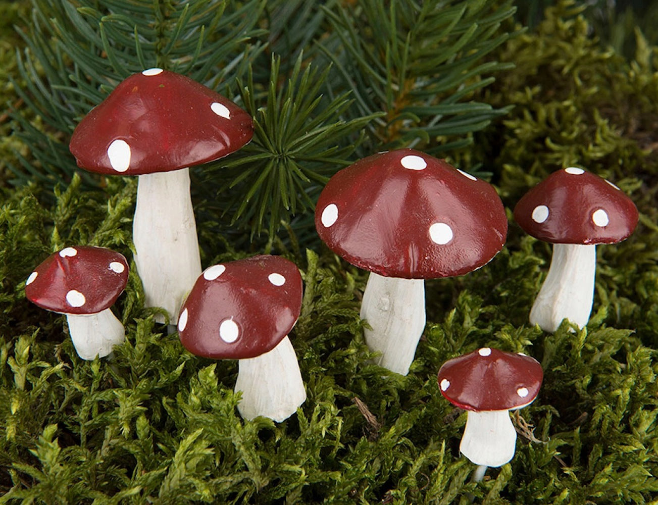 Kompozycja florystyczna z soczyście zielonego mchu i gałązek iglastych, w której umieszczono sześć dekoracyjnych figurek czerwonych muchomorów z białymi kropkami. Grzybki mają różne rozmiary, od bardzo malutkich po nieco większe, i posiadają białe, nierównomierne trzonki, co nadaje im naturalny wygląd.