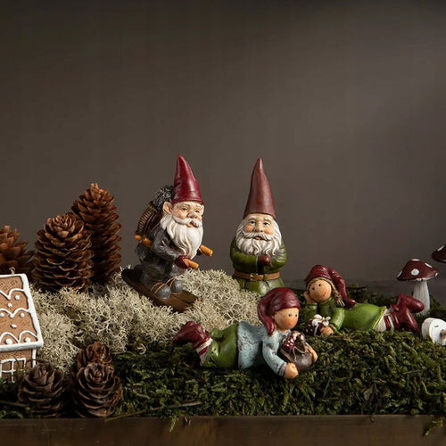 Niezwykle urokliwa, baśniowa diorama inspirowana zimowym lasem. Na naturalnym podłożu z soczyście zielonego oraz siwego mchu (chrobotka) zaaranżowano radosną scenkę z ręcznie malowanymi figurkami. Widzimy tu skrzata szusującego na nartach, stojącego krasnala w zielonym kubraczku oraz dwójkę uśmiechniętych dzieci-skrzatów leżących na brzuchach. Leśny klimat kompozycji dopełniają duże, naturalne szyszki, dekoracyjny muchomor oraz fragment świątecznego domku z piernika. Ciemne, stonowane tło wspaniale podkreśl