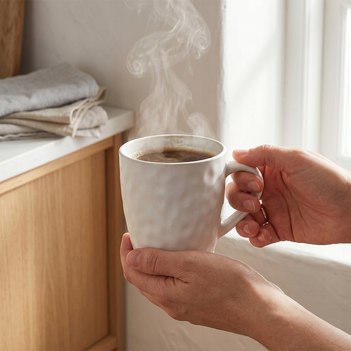 Jasne, lifestylowe ujęcie pełne porannego ciepła. Kadr skupia się na dłoniach delikatnie obejmujących biały, młotkowany kubek z gorącą, parującą kawą. W tle widać fragment jasnej, drewnianej szafki z ułożonymi lnianymi tekstyliami oraz okno, przez które wpada miękkie, naturalne światło. Zdjęcie idealnie oddaje przyjemność płynącą z powolnego, porannego rytuału