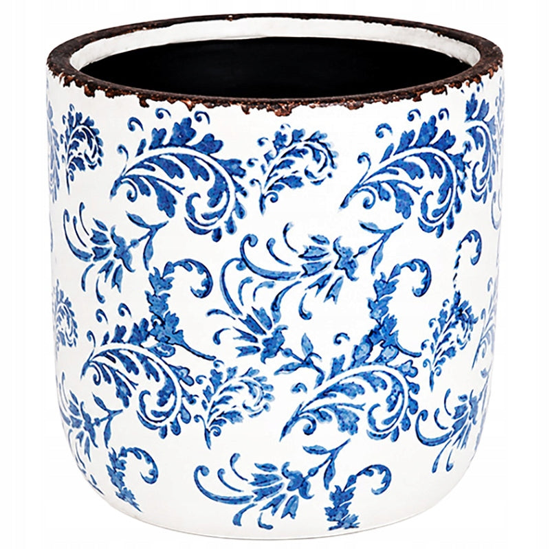 Pusta, cylindryczna donica (osłonka) ceramiczna o błyszczącym wykończeniu. Jej białą powierzchnię w całości pokrywa bogaty, niebieski ornament roślinny, nawiązujący do klasycznej majoliki lub holenderskich wzorów z Delft. Górna krawędź naczynia jest celowo postarzana – wykończona nierównym, ciemnobrązowym rantem imitującym rdzę i otarcia. Zdjęcie produktowe typu packshot wykonane na jednolitym, białym tle.
