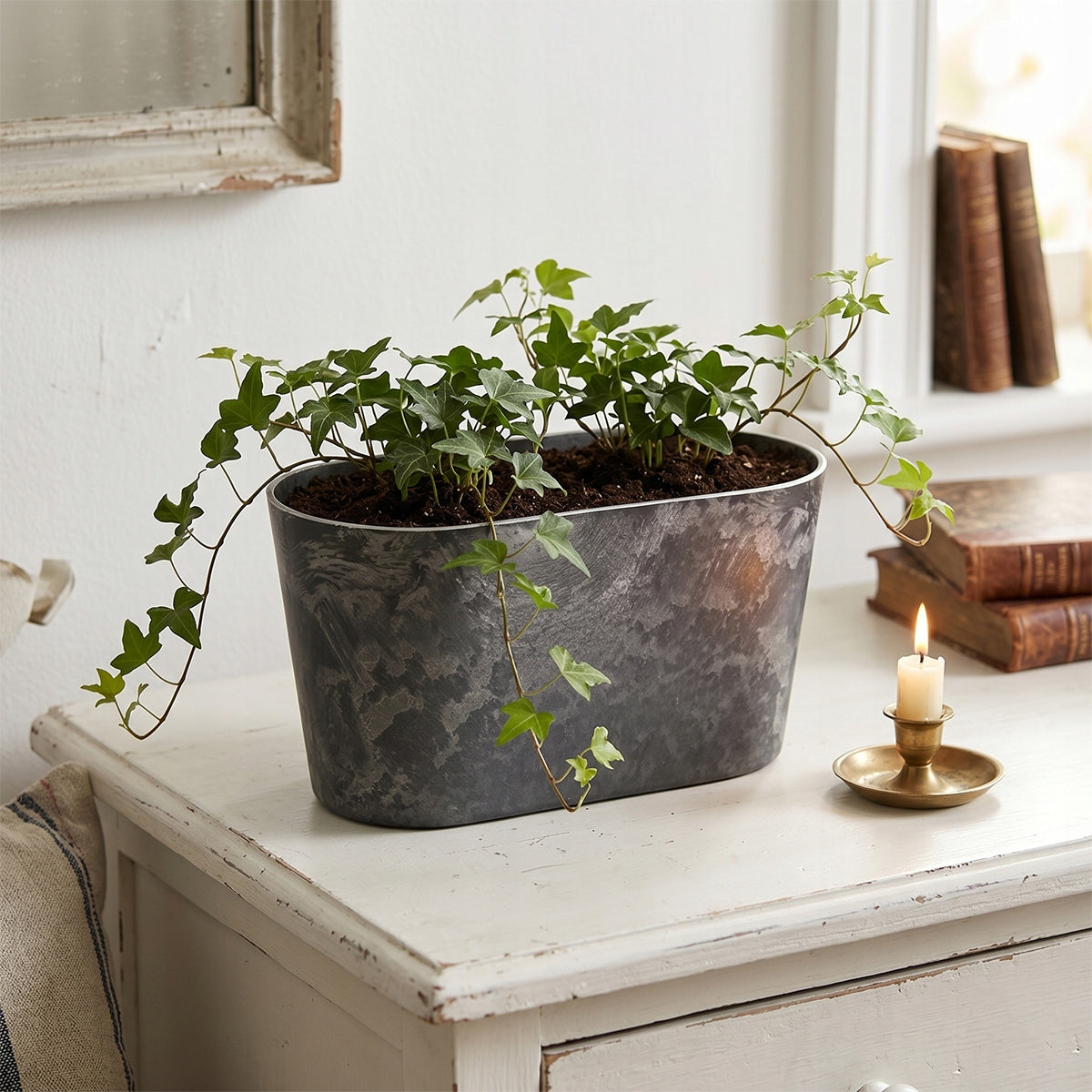 Romantyczna aranżacja w przytulnym stylu vintage lub shabby chic. Podłużna, ciemnoszara donica z pięknie rozrastającym się, zwisającym bluszczem została wyeksponowana na starej, przecieranej, białej komodzie. Obok naczynia stoi mosiężny świecznik z małą, zapaloną świecą oraz zabytkowe książki. Ciepły blask płomienia wspaniale kontrastuje z chłodnym, kamiennym odcieniem donicy.