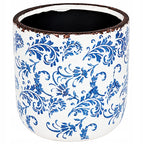 Pusta, okrągła osłonka ceramiczna zaprezentowana na jednolitym, białym tle (zdjęcie typu packshot). Jej zewnętrzną powierzchnię zdobi bogaty, niebieski ornament roślinny przypominający tradycyjną majolikę. Górna krawędź naczynia została celowo postarzona – wykończono ją nierównomiernym, ciemnobrązowym rantem imitującym rdzę i przetarcia. Wnętrze donicy ma kontrastujący, czarny kolor