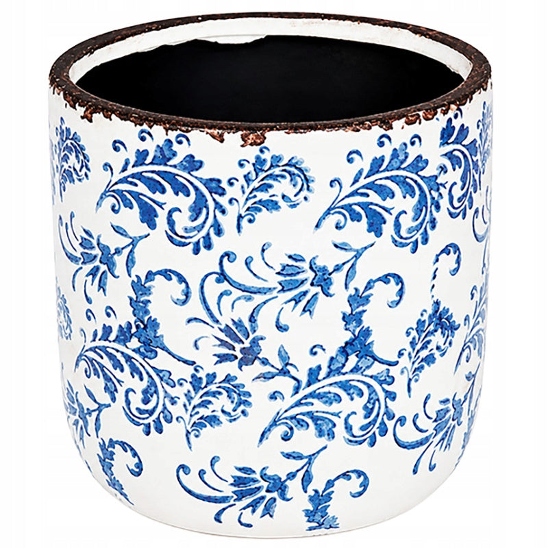 Pusta, okrągła osłonka ceramiczna zaprezentowana na jednolitym, białym tle (zdjęcie typu packshot). Jej zewnętrzną powierzchnię zdobi bogaty, niebieski ornament roślinny przypominający tradycyjną majolikę. Górna krawędź naczynia została celowo postarzona – wykończono ją nierównomiernym, ciemnobrązowym rantem imitującym rdzę i przetarcia. Wnętrze donicy ma kontrastujący, czarny kolor