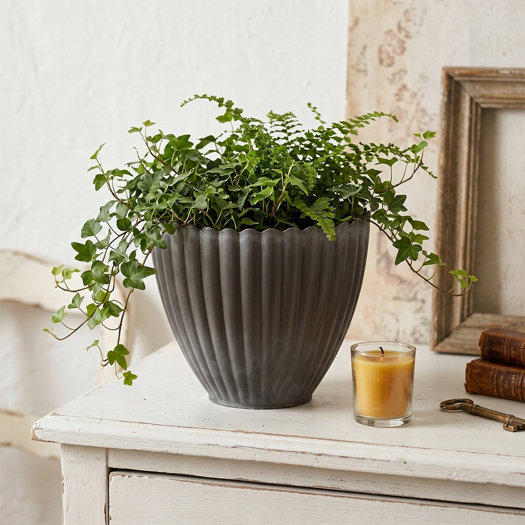 Romantyczna aranżacja w stylu shabby chic. Ciemnoszara, ryflowana donica z bujnie rosnącym, zwisającym bluszczem i drobną paprocią została wyeksponowana na starej, przecieranej, białej komodzie. Obok naczynia stoi zapalona, miodowa świeca w szklanym świeczniku, zabytkowy mosiężny klucz oraz stare książki. Jasne tło nadaje kompozycji przytulny, bardzo nostalgiczny klimat.
