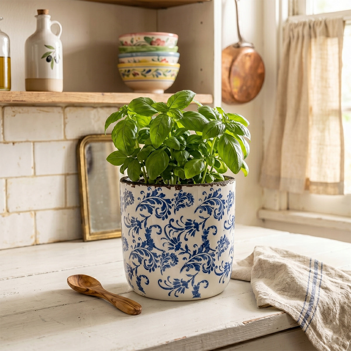 Ceramiczna donica w niebieskie ornamenty wykorzystana jako pojemnik na krzaczek świeżej, pachnącej bazylii. Ustawiona na starym, białym, drewnianym blacie w rustykalnej kuchni. Obok leży mała, drewniana łyżeczka oraz lniana ściereczka z niebieskim paskiem. W przytulnym tle widać białe kafelki, butelkę z oliwą, stosik kolorowych miseczek i miedziany rondel, co tworzy ciepły, sielski klimat