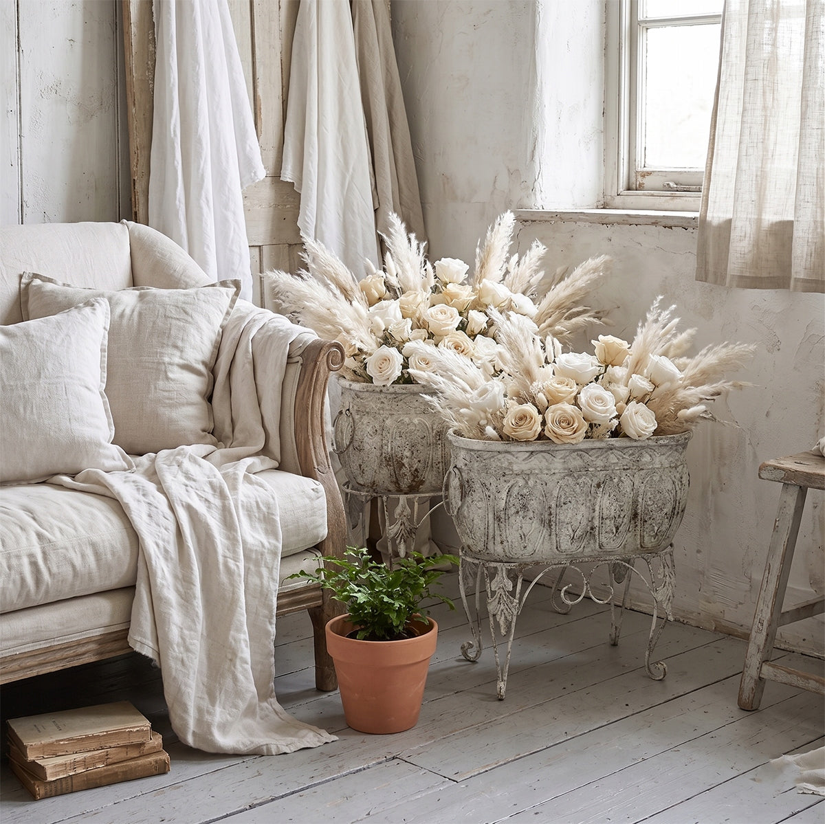 Wnętrze w stylu shabby chic, utrzymane w jasnych, śmietankowych barwach. Dwie podłużne, metalowe donice na stojakach w kolorze przecieranej bieli pełnią rolę okazałych wazonów. Są wypełnione bujnymi kompozycjami z jasnych róż i puszystej trawy pampasowej. Z lewej strony stoi elegancka sofa z narzuconą jasną narzutą i poduszkami. U dołu kompozycję uzupełnia mała, gliniana doniczka z zieloną paprotką oraz stos starych książek na podłodze