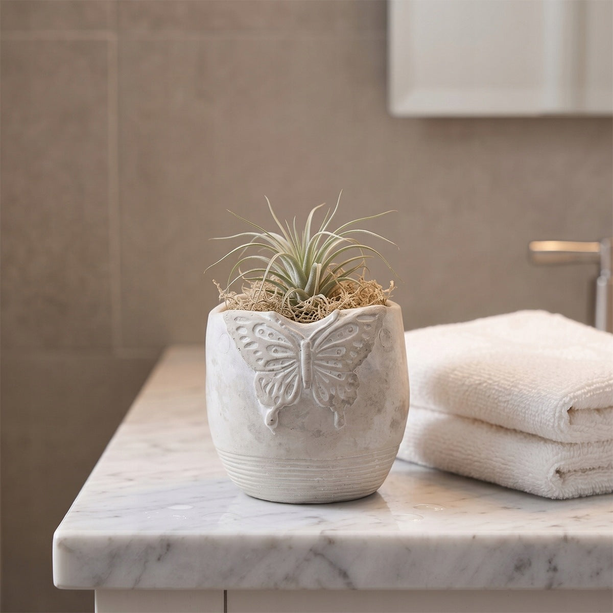 Minimalistyczna, jasna aranżacja łazienkowa w stylu spa. Na jasnym, marmurowym blacie stoi szara, betonowa doniczka z wypukłym dekorem motyla. Wnętrze naczynia wypełnione jest suchym mchem chrobotkiem, z którego wyrasta zielono-srebrzysta oplątwa (roślina powietrzna, Tillandsia). Obok leżą dwa starannie złożone, puszyste białe ręczniki kąpielowe. W lekko rozmytym tle widać fragment srebrnej baterii umywalkowej i gładką ścianę.