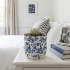 Ceramiczna donica z niebieskim wzorem florystycznym, ustawiona na białej szafce nocnej w jasnej, eleganckiej sypialni. Wewnątrz posadzono estetyczny, zielony sukulent w kształcie rozety. Aranżację uzupełniają leżące obok książki w niebieskich oprawach, ramka ze zdjęciem oraz łóżko z pościelą w paski, co nadaje wnętrzu świeży, lekko marynistyczny klimat w stylu Hamptons.