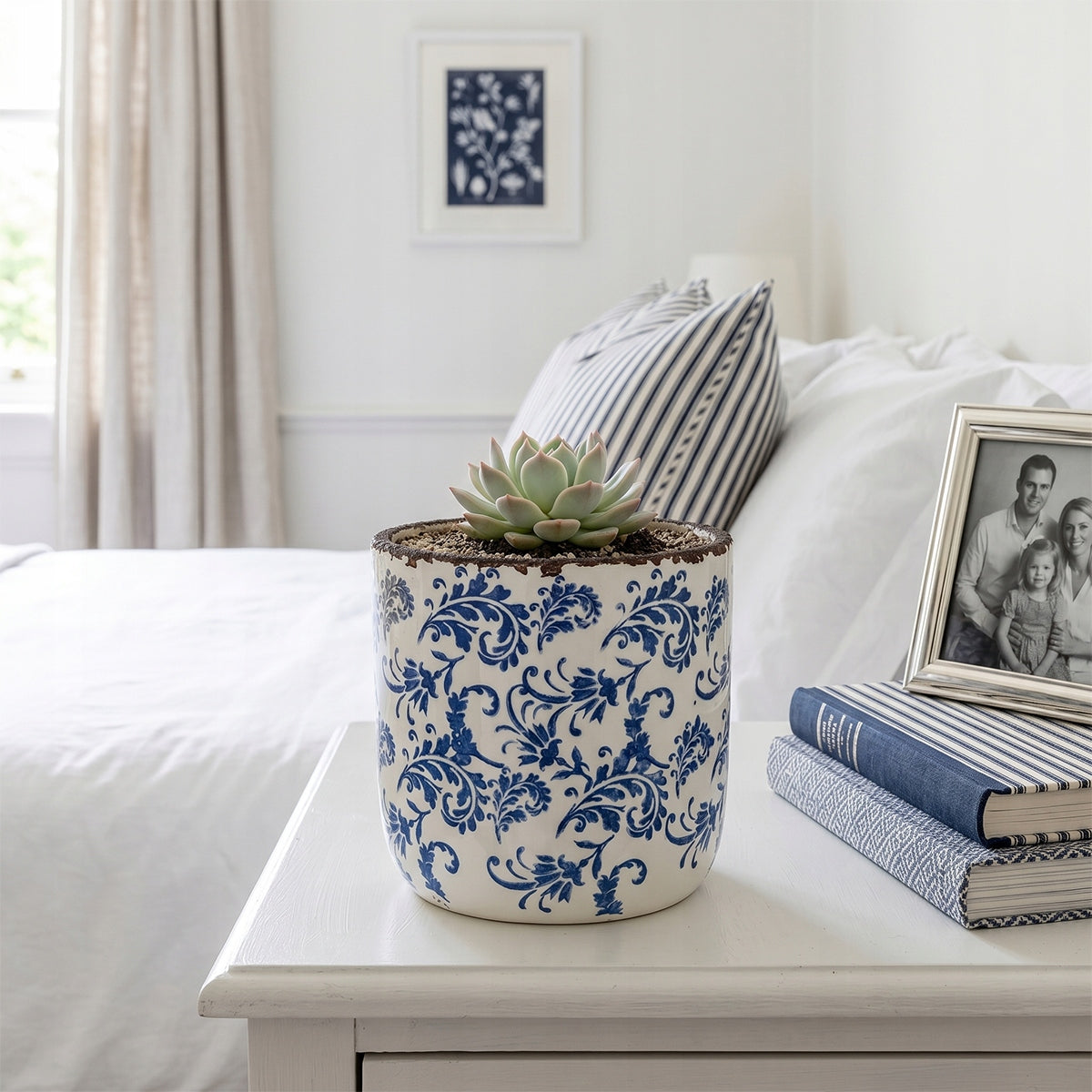 Ceramiczna donica z niebieskim wzorem florystycznym, ustawiona na białej szafce nocnej w jasnej, eleganckiej sypialni. Wewnątrz posadzono estetyczny, zielony sukulent w kształcie rozety. Aranżację uzupełniają leżące obok książki w niebieskich oprawach, ramka ze zdjęciem oraz łóżko z pościelą w paski, co nadaje wnętrzu świeży, lekko marynistyczny klimat w stylu Hamptons.