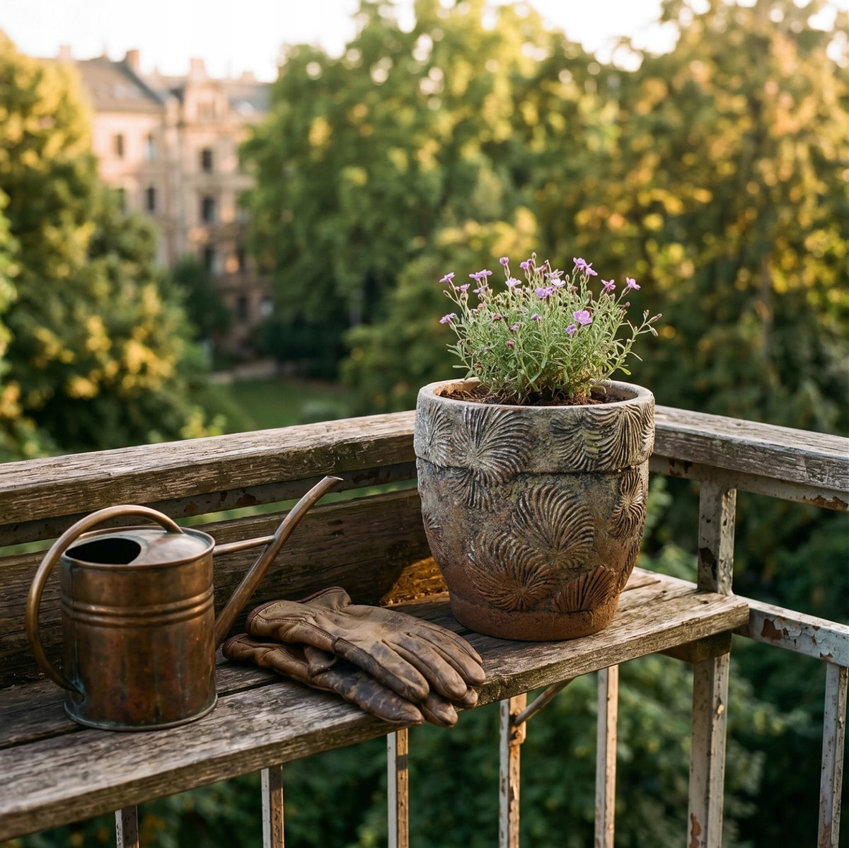 Nastrojowa, letnia aranżacja na balkonie o zachodzie słońca. Na wyblakłej, drewnianej półce przymocowanej do balustrady stoi patynowana doniczka z wytłaczanym wzorem, w której kwitną drobne, różowe kwiatuszki. Tuż obok naczynia leżą znoszone, skórzane rękawice ogrodowe oraz miedziana konewka w stylu vintage. W miękko rozmytym tle widać korony zielonych drzew i fasady miejskich kamienic skąpane w ciepłym świetle.