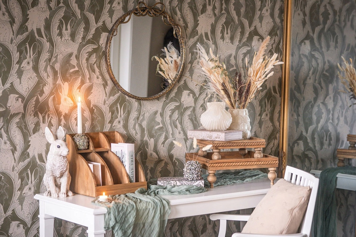 Aranżacja białego biurka w stylu vintage. Na blacie przykrytym szałwiowym bieżnikiem stoi drewniany organizer na dokumenty, figurka białego królika i zapalone świece. Z prawej strony znajdują się dwie piętrowo ułożone, drewniane tace na toczonych nóżkach, które pełnią funkcję podestu dla książek i białego wazonu z suszonymi trawami. Ścianę zdobi zielona tapeta w liście i sylwetki zajęcy, na której wisi okrągłe lustro w ozdobnej ramie.