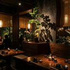 Nastrojowe, eleganckie wnętrze restauracji o azjatyckim charakterze. Na wyeksponowanym, drewnianym podeście stoi duże, sztuczne drzewko bonsai (sosna) o rzeźbiarskim kształcie. Punktowe światło wydobywa głęboką zieleń igieł i rzuca wyrazisty cień rośliny na ciemną, kamienną ścianę za nią. Wokół stoją drewniane stoły z nakryciami, a w tle widać delikatnie oświetlone stoliki i gości.