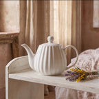 Romantyczna, spokojna kompozycja z białym, prążkowanym dzbankiem ceramicznym. Naczynie ustawiono na starej, białej, drewnianej szafce w jasnej, przytulnej sypialni. Obok dzbanka leży mały bukiecik suszonych ziół: fioletowej lawendy i żółtej mimozy, przewiązany szarym sznurkiem. Światło wpadające przez okno z lnianą zasłoną oraz widoczne w tle łóżko tworzą relaksujący, sielski nastrój