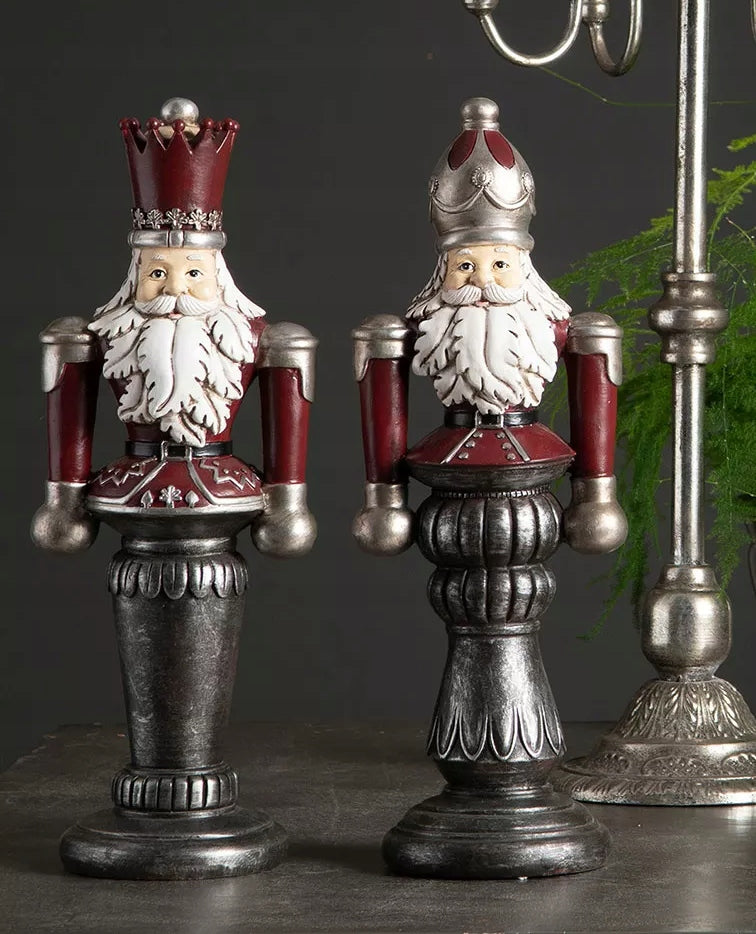 Stylowa kompozycja na ciemnym, matowym blacie. Dwie okazałe figurki Dziadka do orzechów (w bordowych strojach ze srebrnymi detalami i na patynowanych postumentach) zostały wyeksponowane na tle gładkiej, szarej ściany. Z prawej strony kadr dopełnia masywny, klasyczny świecznik w kolorze antycznego srebra oraz subtelna, delikatna gałązka zieleni, tworząc wyrafinowany, nieco mroczny i pałacowy klimat.