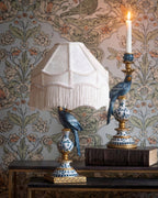 Stylowa aranżacja na ciemnych, metalowych stolikach na tle kwiecistej tapety w stylu vintage. Po lewej stronie stoi lampa z jasnokremowym abażurem z frędzlami; jej podstawa to figura niebieskiej papugi siedzącej na ceramicznym, obłym elemencie w niebiesko-białe wzory ze złotymi akcentami. Po prawej stronie widoczny jest pasujący, wysoki świecznik z identycznym motywem niebieskiej papugi i zapaloną białą świecą, ustawiony na starej, grubej książce.