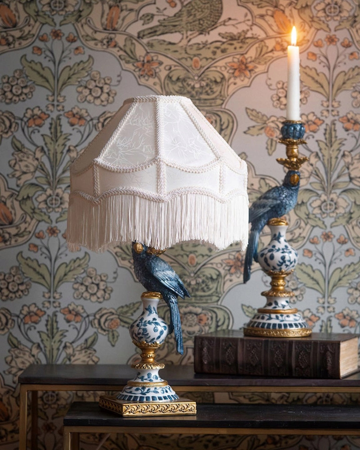Stylowa aranżacja na ciemnych, metalowych stolikach na tle kwiecistej tapety w stylu vintage. Po lewej stronie stoi lampa z jasnokremowym abażurem z frędzlami; jej podstawa to figura niebieskiej papugi siedzącej na ceramicznym, obłym elemencie w niebiesko-białe wzory ze złotymi akcentami. Po prawej stronie widoczny jest pasujący, wysoki świecznik z identycznym motywem niebieskiej papugi i zapaloną białą świecą, ustawiony na starej, grubej książce.