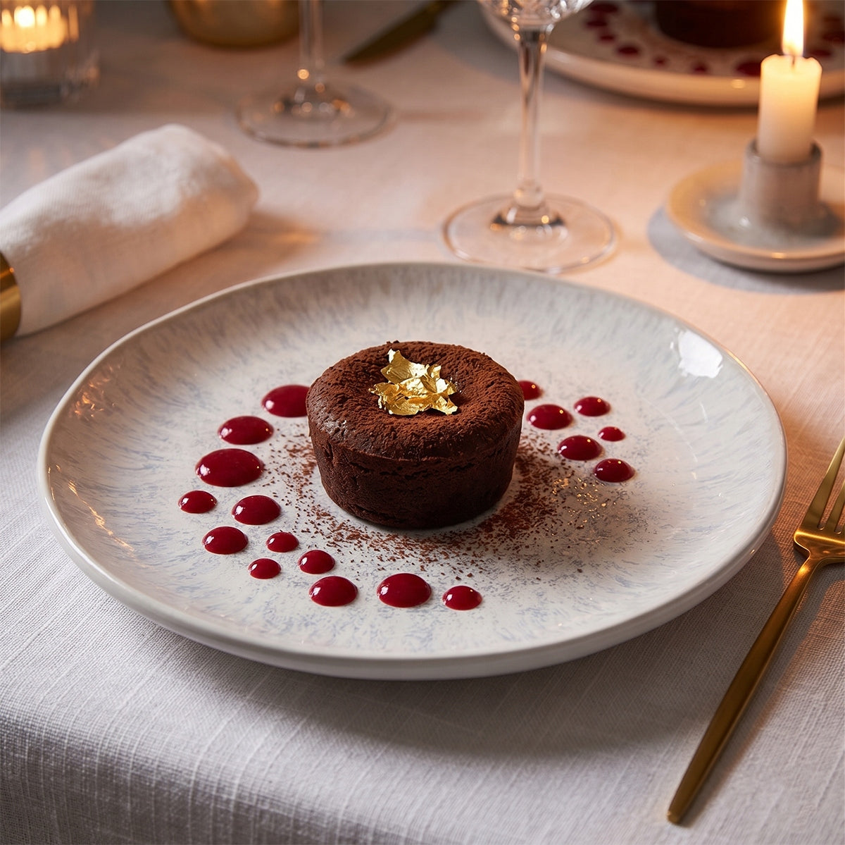 Wykwintna, wieczorna kompozycja kulinarna w stylu fine dining. Na jasnym talerzu pięknie kontrastuje ciemne, czekoladowe ciastko (fondant) oprószone kakao i dumnie zwieńczone jadalnym złotem. Deser otaczają artystycznie ułożone krople intensywnie czerwonego sosu owocowego. Złote sztućce, blask świec, biały obrus i eleganckie szkło w tle tworzą luksusową, romantyczną atmosferę