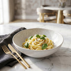Elegancka kompozycja kulinarna zaserwowana na jasnym, marmurowym blacie. W głębokiej, ceramicznej misce o subtelnym, nakrapianym wzorze podano apetyczny makaron (tagliatelle) posypany serem i udekorowany listkami świeżej bazylii. Obok leży ciemnoszara, lniana serwetka oraz stylowe, złote sztućce. Rozmyte tło z jasnym wnętrzem i welurowymi krzesłami nadaje scenie luksusowy, restauracyjny charakter