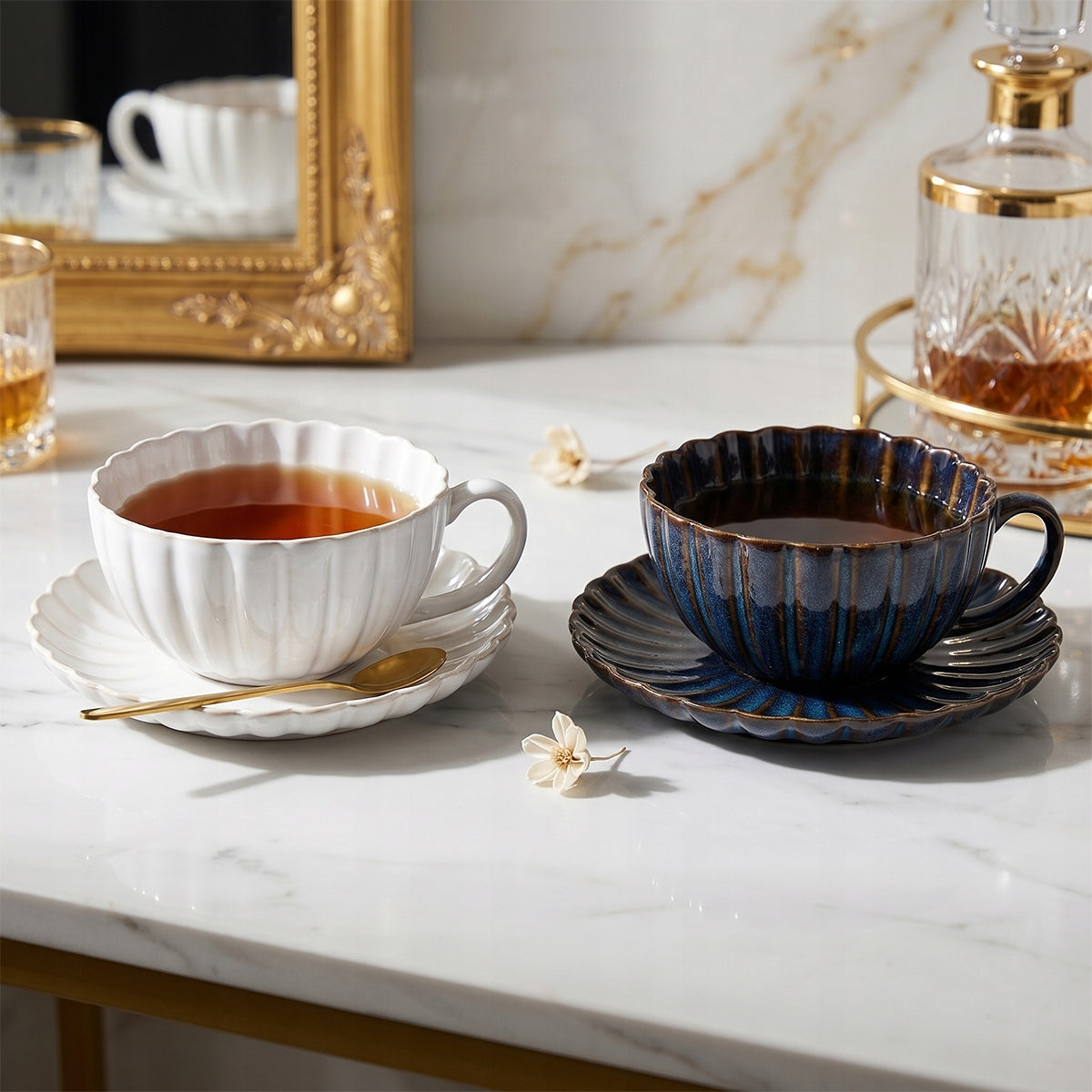 Wykwintna i elegancka kompozycja z wykorzystaniem ceramicznych filiżanek. Naczynia z zaparzoną herbatą stoją na jasnym, marmurowym blacie. Luksusowy charakter aranżacji podkreślają złote akcenty: mała łyżeczka, ozdobna rama lustra w tle oraz taca z kryształową karafką. Delikatne, drobne kwiatki leżące na stole dodają całości subtelności i romantycznego, wysublimowanego nastroju