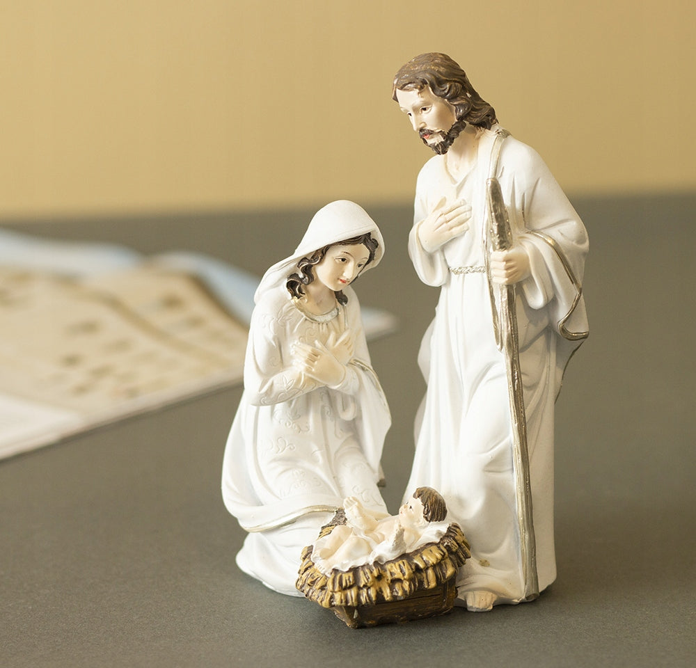 Dekoracyjna figurka Świętej Rodziny (Maryja, Józef i mały Jezus w żłóbku) wyeksponowana na gładkim, szarym blacie. Postacie w jasnych szatach ze złotymi detalami są przedstawione w tradycyjnej scenie narodzenia Jezusa. Tło jest minimalistyczne, utrzymane w ciepłych, pastelowych barwach z delikatnym rozmyciem.