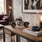 Czarno-srebrny, metalowy model motocykla wyeksponowany na drewnianym blacie industrialnej konsoli z metalową ramą. Obok leży stos książek o tematyce designu, niebieska, ceramiczna popielniczka oraz nowoczesna lampa ze szklanym kloszem. W tle znajduje się surowa, betonowa ściana z czarno-białym obrazem, fioletowa kanapa oraz duża lampa na statywie, tworzące spójną aranżację w stylu loftowym.