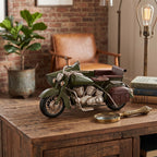 Zielony, metalowy model motocykla z wózkiem bocznym wyeksponowany na rustykalnej, drewnianej konsoli. Obok figurki leży ozdobna, mosiężna lupa retro oraz stos książek. W przytulnym tle w stylu loftowym widać ścianę z czerwonej cegły, brązowy skórzany fotel, dużą roślinę doniczkową w metalowej osłonce oraz stojącą lampę z żarówką Edisona.