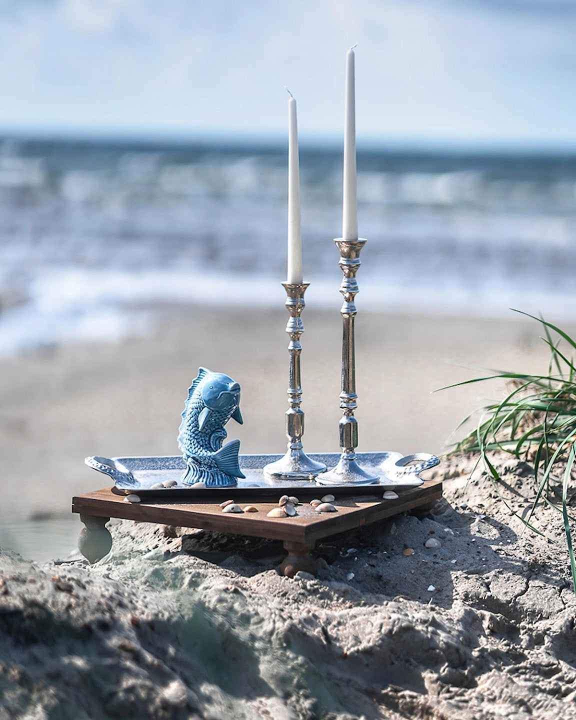 Błękitna figurka ryby ustawiona na ozdobnej, srebrnej tacy, która spoczywa na małym, drewnianym stoliku bezpośrednio na plaży. Na tacy znajdują się również dwa wysokie, klasyczne świeczniki ze srebra z długimi, białymi świecami. Dookoła rozsypane są drobne muszelki, a w tle widać rozmyty krajobraz z falami morza, idealnie oddający wakacyjny, nadmorski klimat.