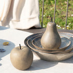 Aranżacja na świeżym powietrzu, na stole przykrytym jasnym obrusem. Kompozycję tworzą ceramiczne figurki dekoracyjne imitujące ciemne drewno z czarnymi ogonkami. Na pierwszym planie leży jabłko, a tuż za nim, w rzeźbionej, ceramicznej misie, stoi podobna figurka w kształcie gruszki. W tle widać krzesło z narzuconym kocem oraz rozświetlony słońcem trawnik.