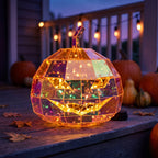 Wieczorna dekoracja tarasu na Halloween. Na drewnianych deskach stoi duża, świecąca dynia o geometrycznej, wielościennej strukturze z holograficzną powłoką, która mieni się w odcieniach pomarańczu i fioletu. Lampa ma wycięte groźne oczy i uśmiech, a wewnątrz niej widać ciepłe światełka. W tle widać prawdziwe dynie oraz zawieszone na balustradzie girlandy świetlne.