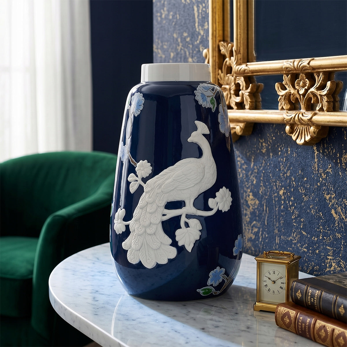 Pusty, granatowy wazon ceramiczny z wyraźnym, białym reliefem pawia, wyeksponowany na okrągłym stoliku z jasnym, marmurowym blatem. Obok niego leżą stare książki w skórzanych oprawach oraz mały, mosiężny zegar. W głębi widać elegancką, ciemnoniebieską ścianę ze złotymi przetarciami, ozdobną, złotą ramę lustra oraz szmaragdowozielony, welurowy fotel, co tworzy luksusową aranżację w stylu glamour.