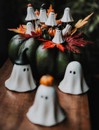 Nastrojowa, mroczna kompozycja na Halloween. Na ciemnym, drewnianym blacie stoi duża, ciemnozielona dynia pełniąca funkcję bazy stroika. Wyrastają z niej sztuczne, jesienne liście w intensywnych odcieniach czerwieni i żółci, małe dynie oraz liczne piki dekoracyjne w kształcie białych duszków (część z nich ma czarne kapelusze lub dynie na głowach). Na pierwszym, mocno rozmytym planie stoją bezpośrednio na blacie trzy dodatkowe figurki duszków.