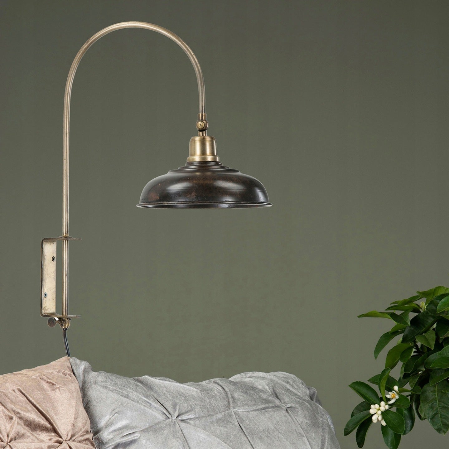 Kinkiet ścienny w stylu industrialnym/loftowym zamontowany na tle gładkiej, ciemnooliwkowej ściany. Lampa posiada mosiężne, gięte ramię i klosz z ciemnego, postarzanego metalu, który oświetla strefę wypoczynkową. Poniżej lampy widać krawędź kanapy z welurowymi, pikowanymi poduszkami w kolorze szarym i pudrowego różu, a z prawej strony kadr zamyka fragment zielonej rośliny.